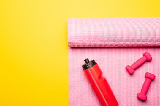 top view of red sports bottle, pink fitness mat and dumbbells on yellow background