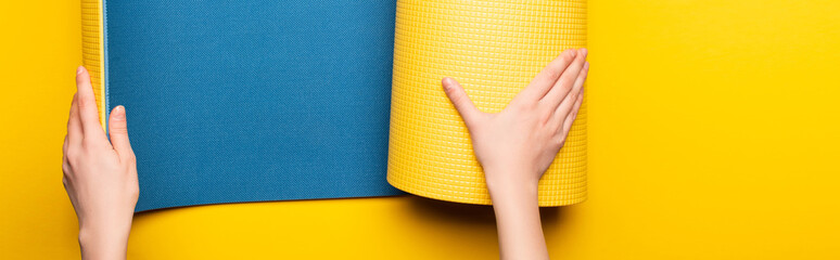 partial view of woman rolling out blue fitness mat on yellow background, panoramic shot