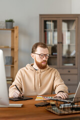 Fototapeta premium Serious young bearded man in eyeglasses drawing design sketch on graphics tablet and editing it on laptop