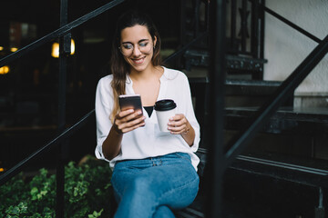 Smiling caucasian hipster girl smiling while checking messages and notification on smartphone sitting on urban stairs, positive young woman watching videos and vlogs on modern mobile phone