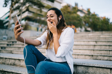 Emotional caucasian woman in earphones having video call on mobile phone surprised with news while taking, happy female millennial blogger amamzen with social networks updates using smartphone