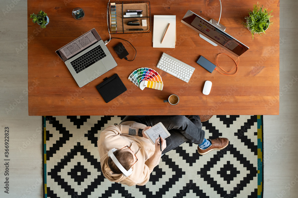 Wall mural above view of modern hipster designer in headphones using digital tablet while working on scheme in 