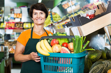 Mature saleswoman is demonstraiting basket with variety goods in greengrocery