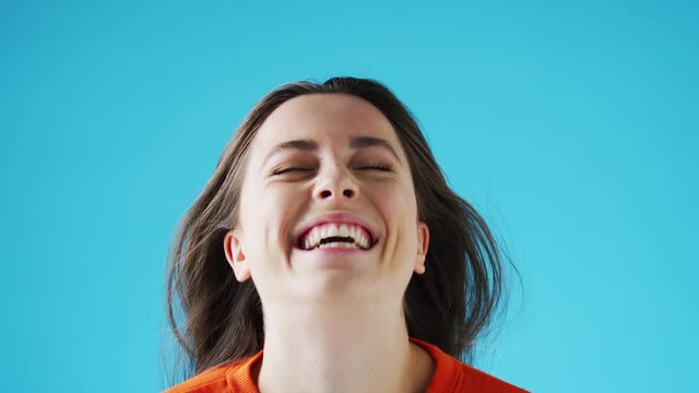Studio portrait of smiling young woman with long hair throwing her head back into frame and laughing on blue background - shot in slow motion