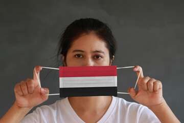 Naklejka premium A woman in white shirt with Yemen flag on hygienic mask in her hand and lifted up the front face on dark grey background. Tiny Particle or virus corona or Covid 19 protection.