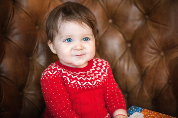 A little girl with blue eyes in a red dress sits in a brown leather armchair, close-up portrait.