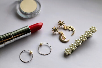 Lipstick, eyeshadow, gold rings, earrings and pearl barrette on pale lilac background. Selective focus.