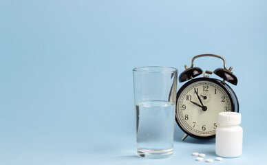 Jar of pills, water and a clock on a blue background. Time to take pills. Insomnia. Copy space.