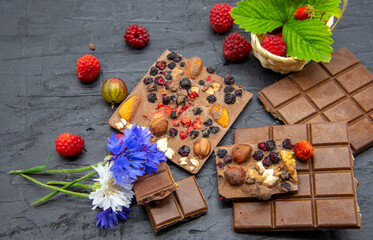 A few slices of chocolate, berries, flowers, and a basket of strawberry leaves on a black slate background. Copy space for text, design.