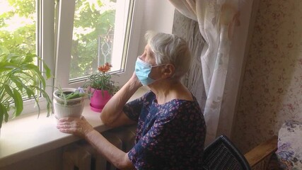 The Covid-19, health, safety and pandemic concept - senior old lonely woman wearing protective medical mask sitting near the window at home for protection from virus