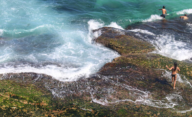 Coastal rocky outcrop with algae / moss, woman walking on slippery rocks, guys jumping into the ocean. Sydney