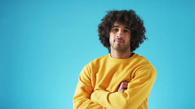 Studio portrait of confident young man looking into camera with neutral expression and folded arms against blue background - shot in slow motion