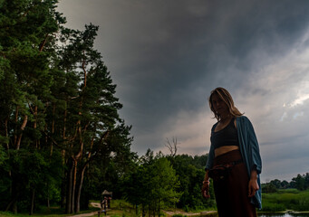 Fototapeta na wymiar Young woman in full growth on background of pine trees and an approaching thunderstorm