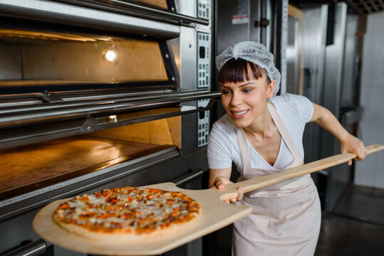 Young Caucasian Woman Baker Is Holding A Wood Peel With Fresh Pizza Near An Oven At A Baking Manufacture Factory.