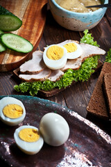 assembling sandwich with meat, dark bread, fresh green salad, eggs and cucumbers on rustic wooden table surface
