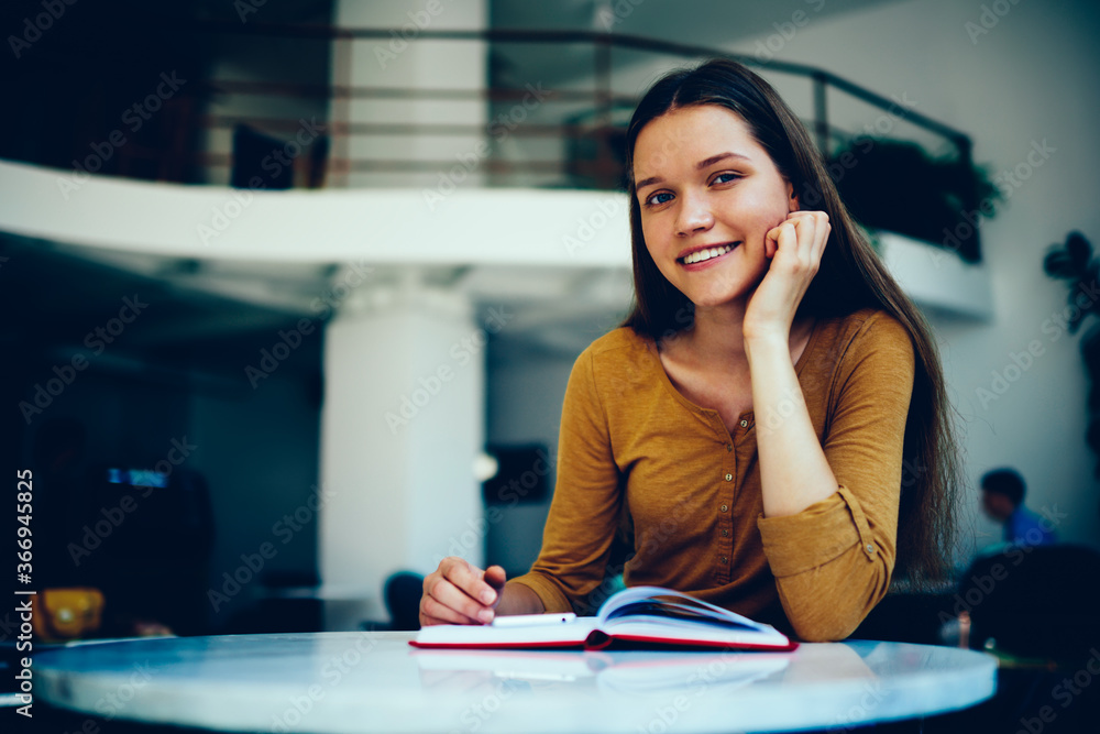 Aufkleber portrait of charming student looking at camera while reading interesting book preparing for exams in
