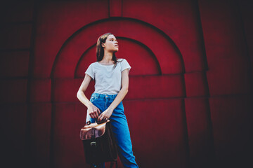 Fototapeta na wymiar Beautiful hipster girl in casual outfit posing on red urban wall background carrying trendy leather bag,attractive young female traveler spending time outdoors standing on street with backpack