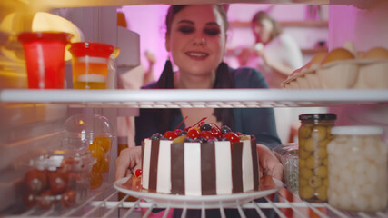 Young curvy woman taking cake from fridge with friends having fun at home party on background