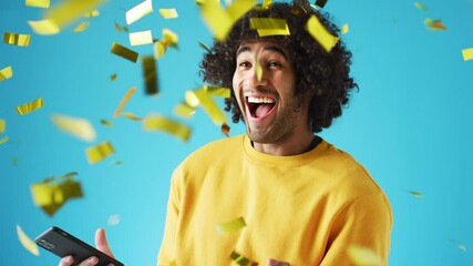 Celebrating young man with mobile phone winning prize and showered with gold confetti in studio - shot in slow motion