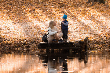 Mother and son are sitting on a wooden pier near the lake in the autumn park