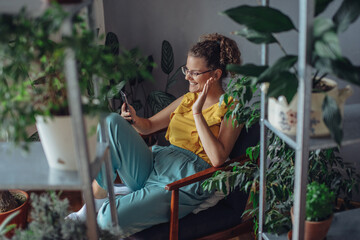 Young woman using mobile phone. Young florist in eyeglasses sitting on armchair and using mobile phone in the room with plants.