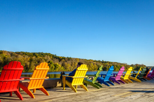 Man relaxing in a row of brightly colored adirondack chairs along waterfront in fall/autumn