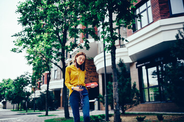 Fototapeta na wymiar Pondering cute hipster girl checking notification on smartphone connected to 4G internet while strolling at urban setting.Pensive female dressed in yellow blouse and jeans listening to favorite radio