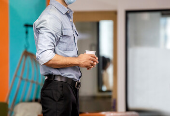 Young man worker wear face mask and holding paper glass in modern office