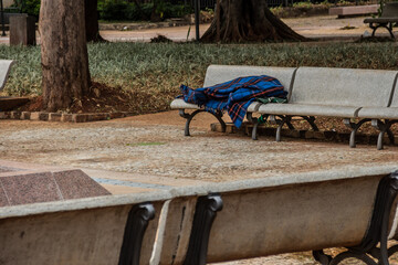 Pessoa sem teto, embrulhado em coberta, dormindo em banco de praça.