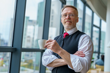 Fototapeta premium Senior businessman wears white shirt and drinking coffee near big window with city view . Photo from the side.