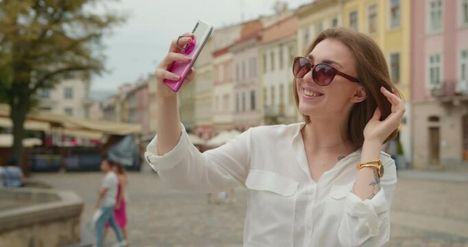 A girl is taking a selfie when standing in the central square. She is smiling and is happy. She is wearing sunglasses. The people are walking in the background. 4K