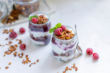 Homemade organic granola or muesli with natural yogurt, fresh raspberries and mint leaf in a jars, white table near window. Healthy beautiful background, sunny light. Selective focus, copy space.