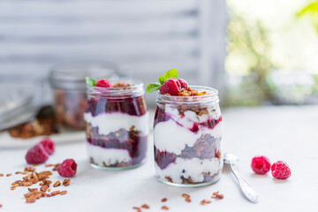 Homemade organic granola or muesli with natural yogurt, fresh raspberries and mint leaf in a jars, white table near window. Healthy beautiful background, sunny light. Selective focus, copy space.