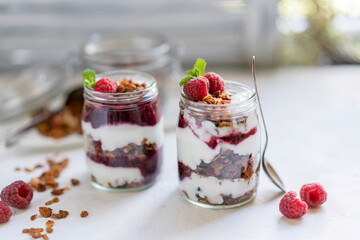 Homemade organic granola or muesli with natural yogurt, fresh raspberries and mint leaf in a jars, white table near window. Healthy beautiful background, sunny light. Selective focus, copy space.