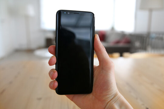 A male hand holding a brand new, very modern, state of the art mobile phone. The touch screen is black with reflections on it. An image on an interior design background with a visible wood table.
