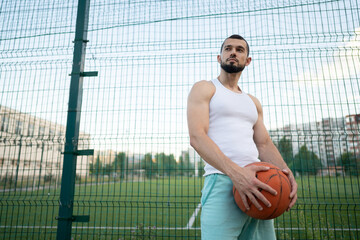 Man stands near a fence on the street, holding a basketball in his hand