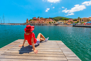 Tourist woman sunbathing on the jetty in Rio Marina harbor of Elba island in red suit. Lifestyle tourist girl on holiday travel in Italy, Europe. Rio Marina skyline cityscape on background