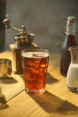 A cup of ice cold brew coffee in yellow light on wood table. Milk bottle and coffee grinder in the background. Side view. Made from Arabica bean