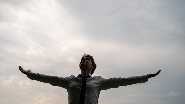Young businessman standing under cloudy grey sky aith his arms spread widely