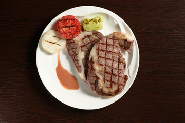 Two fried marble beef steaks with grilled vegetables of tomato, onion and bell pepper on a round plate on a wooden table. Top view. Photos for restaurant and cafe menus