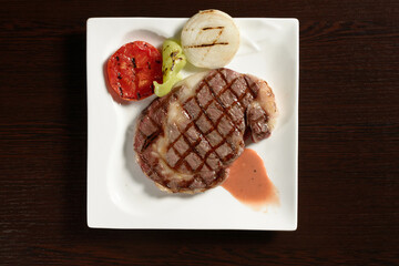 Fried marble beef steaks with grilled vegetables of tomato, onion and bell pepper on a square plate on a wooden table. Top view. Photos for restaurant and cafe menus