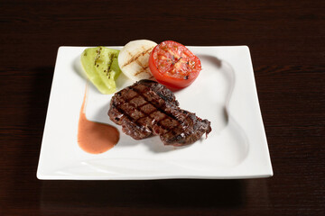 Roasted meat of marble beef steaks with grilled vegetables of tomato, onion and bell pepper on a square plate on a wooden table. Photos for restaurant and cafe menus