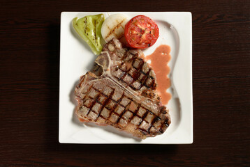 T-bone grilled beef steak with grilled vegetables of tomato, onion and bell pepper on a square plate on a wooden table. Top view. Photos for restaurant and cafe menus