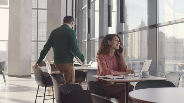 Pan shot of young curly-haired businesswoman sitting at desk in modern office and talking on telephone while middle-aged businessman coming and sitting at the next table