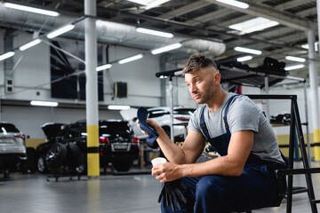 Fototapeta na wymiar tired technician in overalls holding cap and disposable cup while sitting near cars in service station