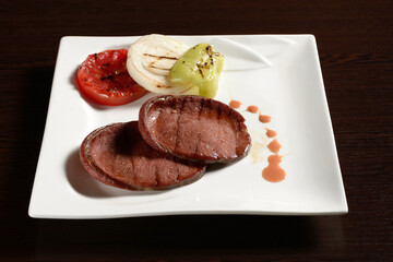 Grilled slices of beef sausage with fried vegetables of tomato, onion and bell pepper on a square plate on a wooden table. Photos for restaurant and cafe menus