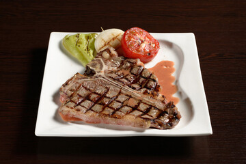 Concept of cooking meat. Roasted t-bon beef steak with grilled vegetables of tomato, onion and bell pepper on a square plate on a wooden table. Photos for restaurant and cafe menus
