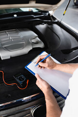 Cropped view of auto mechanic writing on clipboard near open hood of car with multimeter at service...