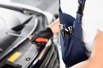 selective focus of mechanic in overalls with metallic tools in pocket standing near in workshop
