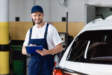 happy mechanic in uniform holding clipboard near modern car